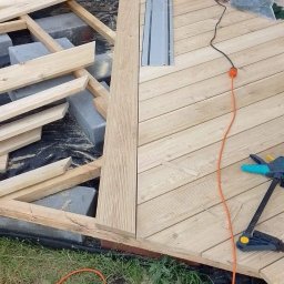 Sztuka Drewna Tworzymy z Pasją - Montaż drewnianego tarasu: fragment ułożonych desek, widoczna konstrukcja nośna z bloczków betonowych, narzędzia pracy - piła tarczowa z szyną prowadzącą, zacisk stolarski i przewód zasilający.