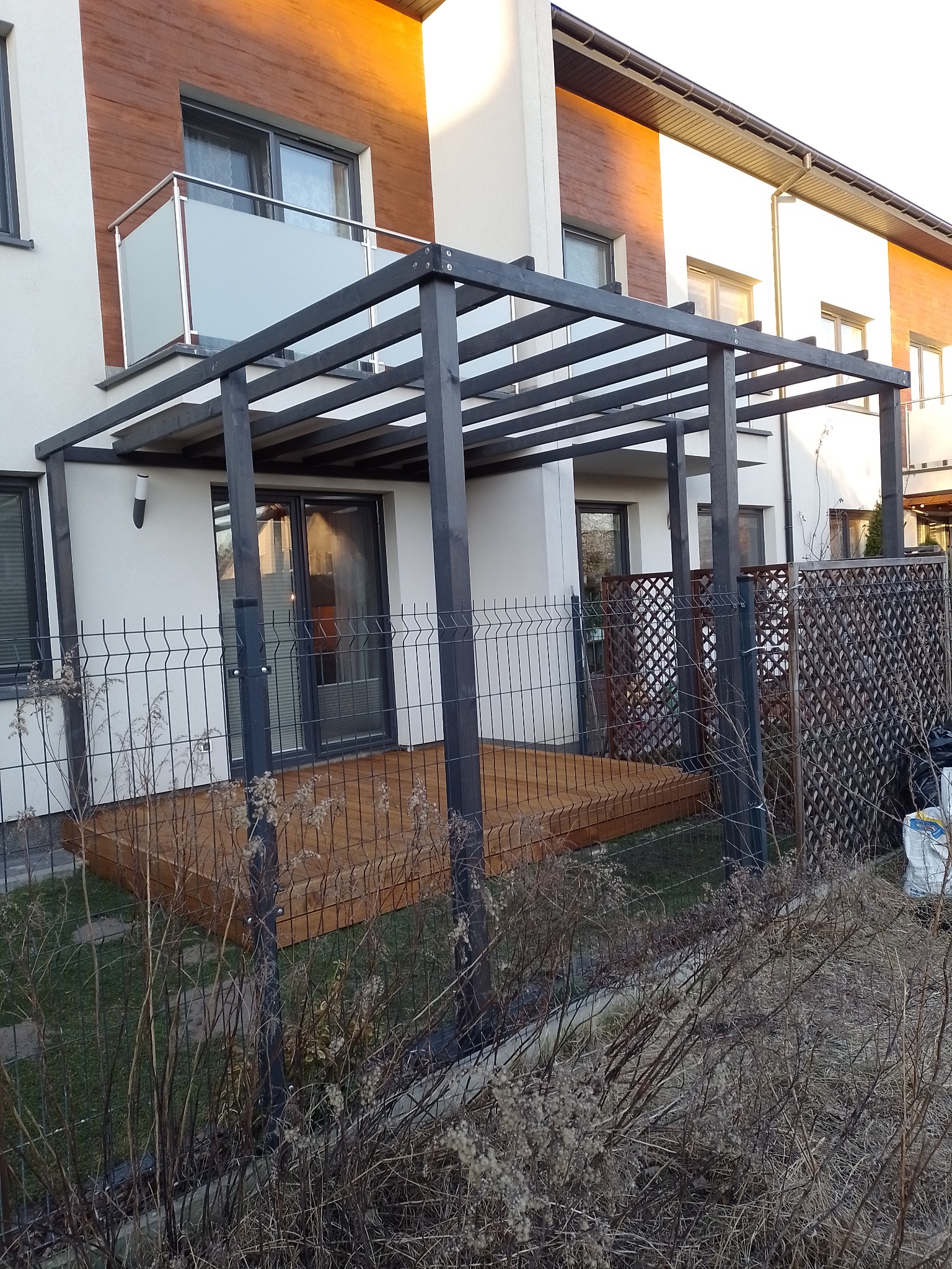 Ciemnoszara pergola z drewnianym zadaszeniem nad drewnianym tarasem przylegającym do nowoczesnego domu z balkonem, widok zza metalowego ogrodzenia.