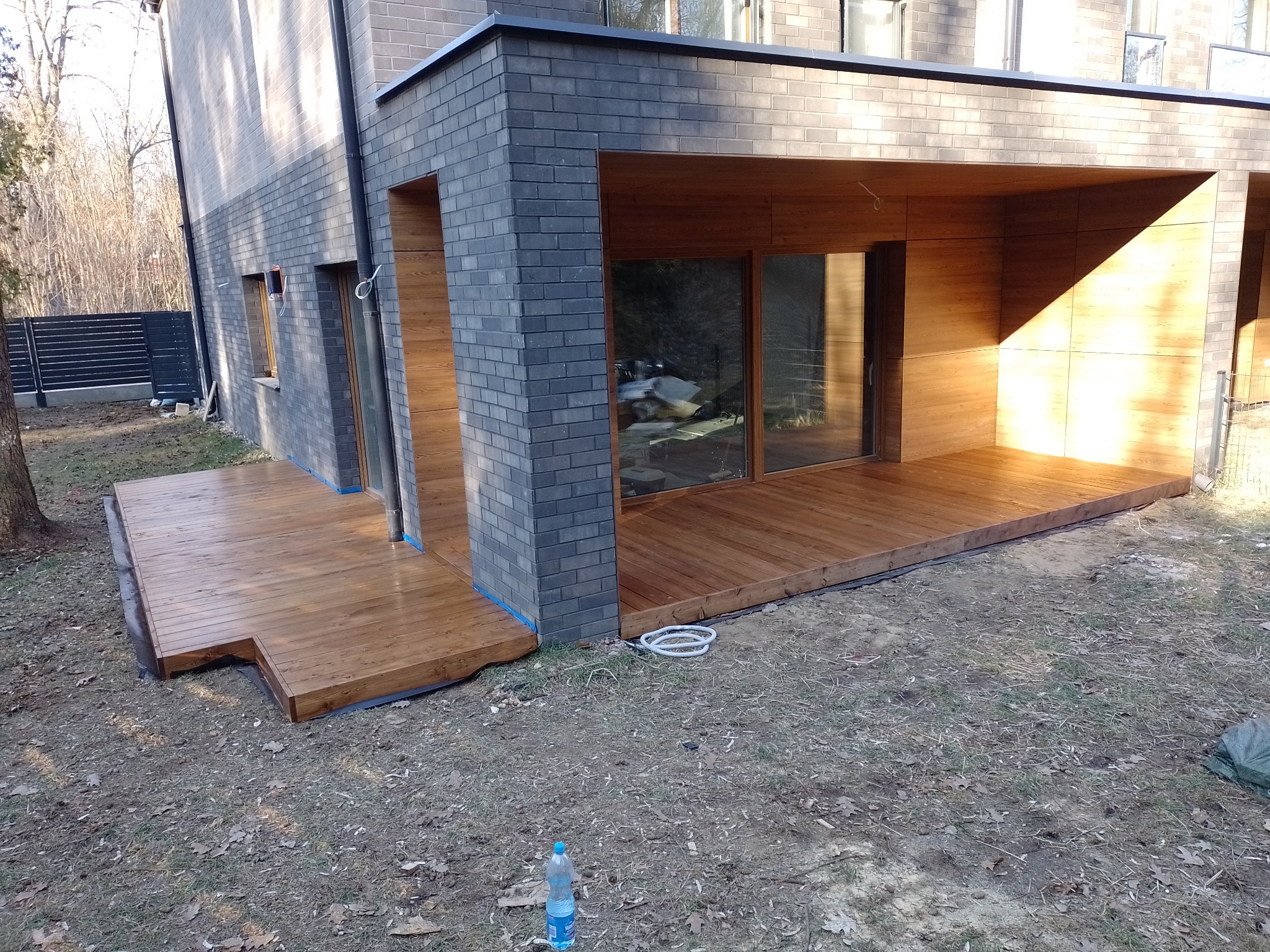 Nowoczesny dom z szarej cegły z przylegającym drewnianym tarasem i zadaszeniem, widoczne niebieskie taśmy malarskie, butelka wody i zwinięty biały wąż na ziemi.