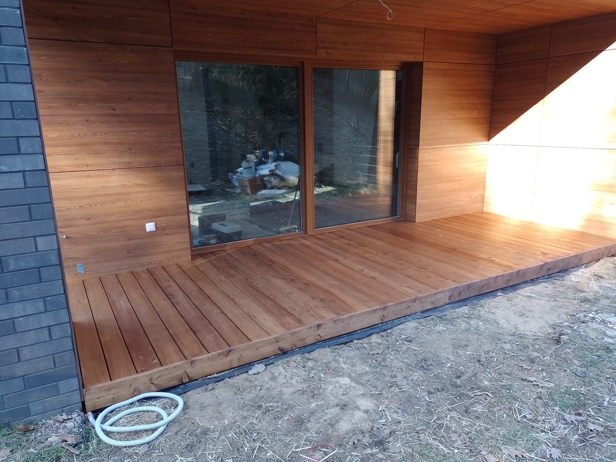 Nowoczesny taras drewniany przylegający do domu z drewnianą elewacją, widoczny fragment ściany z szarej cegły oraz biały wąż ogrodowy na ziemi.