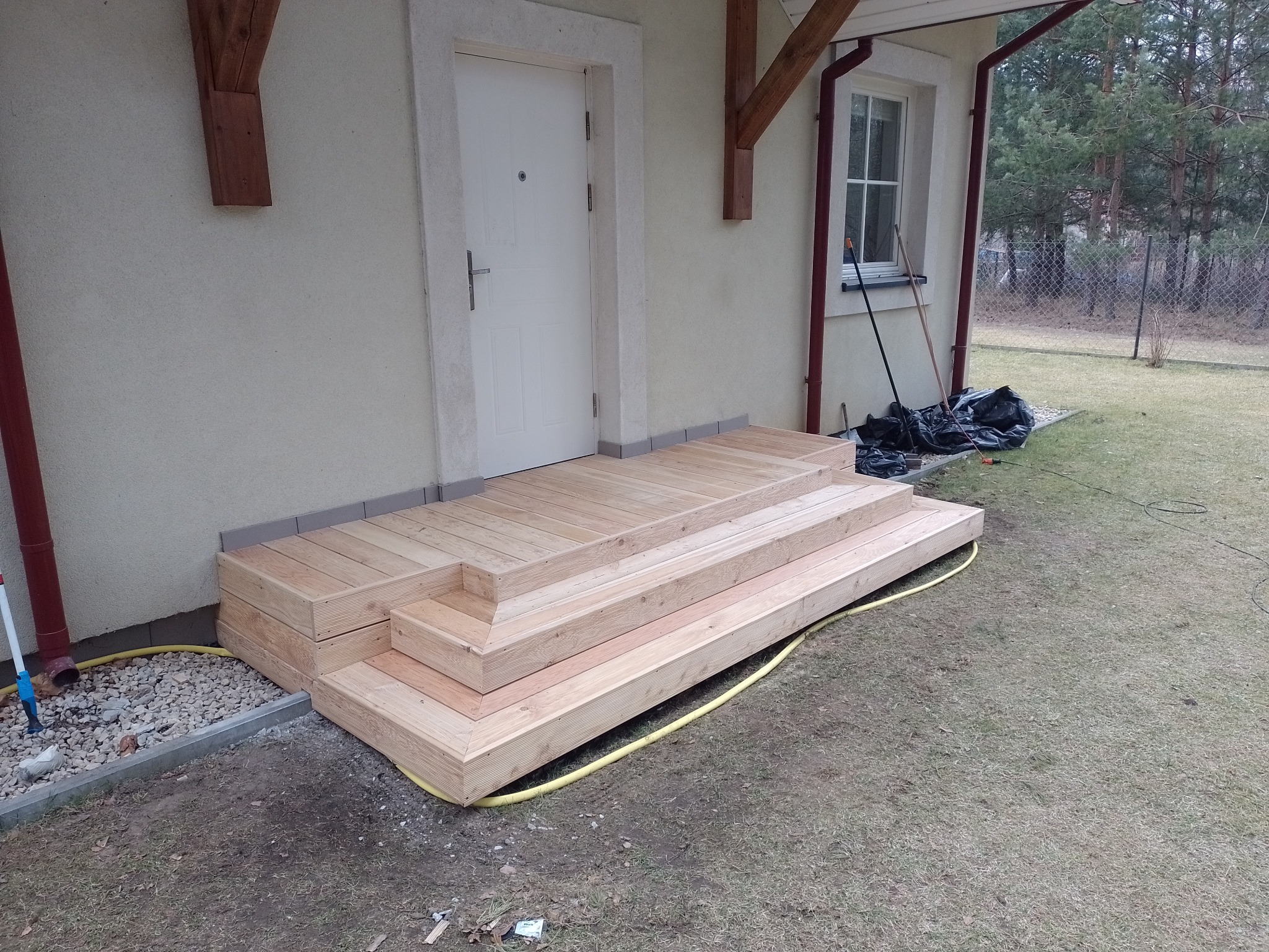 Schody wejściowe z jasnego drewna prowadzące do drzwi domu, z widoczną żółtą rurą i narzędziami w tle.