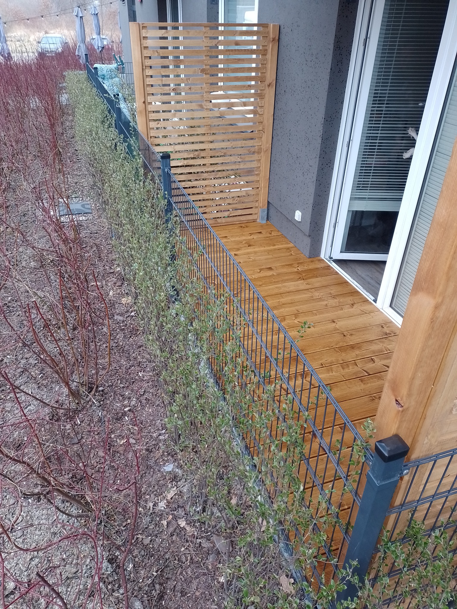 Balkon z drewnianą podłogą i ażurową osłoną, widok z góry na metalową balustradę i krzewy