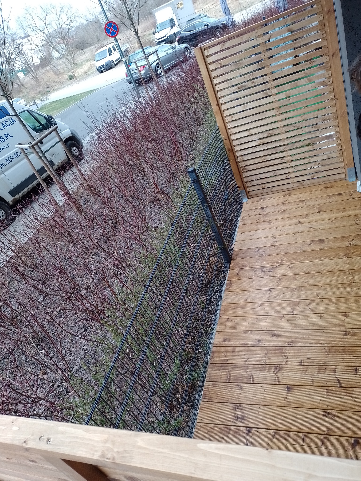 Drewniany taras z ażurową osłoną i czarnym metalowym ogrodzeniem, widok z góry na ulicę i zaparkowane samochody.