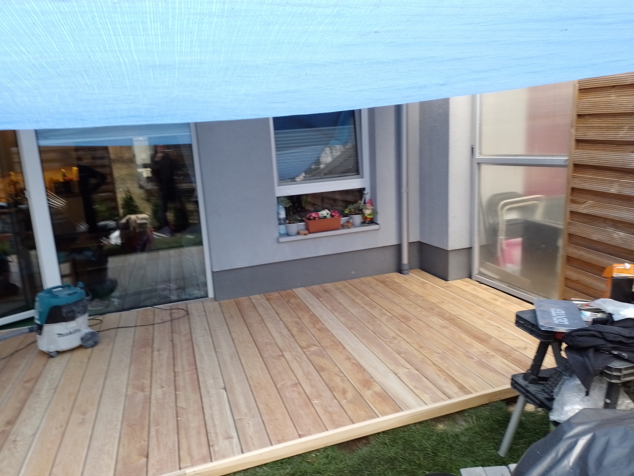 Nowa, jasna drewniana podłoga tarasu przylegająca do domu, częściowo osłonięta niebieską plandeką, odkurzacz budowlany i stolik warsztatowy z narzędziami w tle.