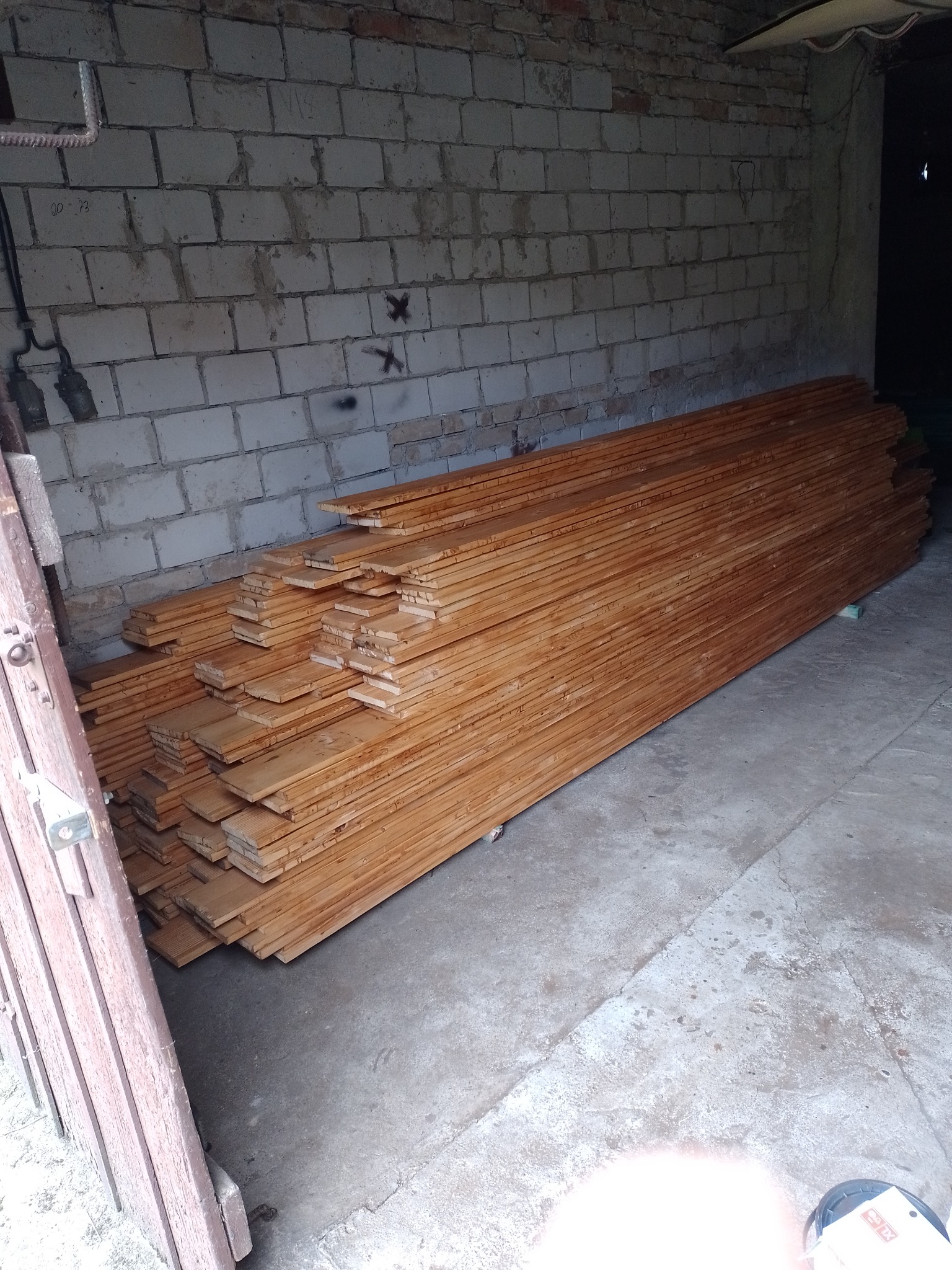 Stos surowych desek z jasnego drewna ułożony w pomieszczeniu z ceglaną ścianą w tle, widoczne ślady po naprawach na ścianie.