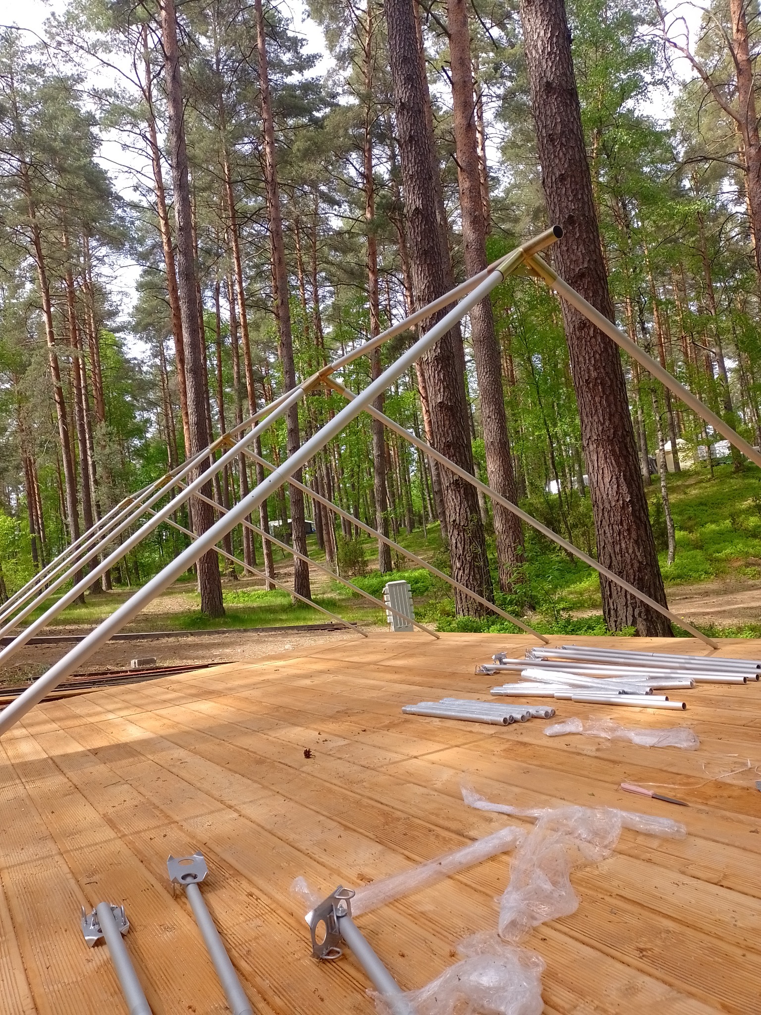 Montaż metalowej konstrukcji w lesie na drewnianym podłożu, widoczne elementy mocujące i rury, częściowo z folią ochronną.