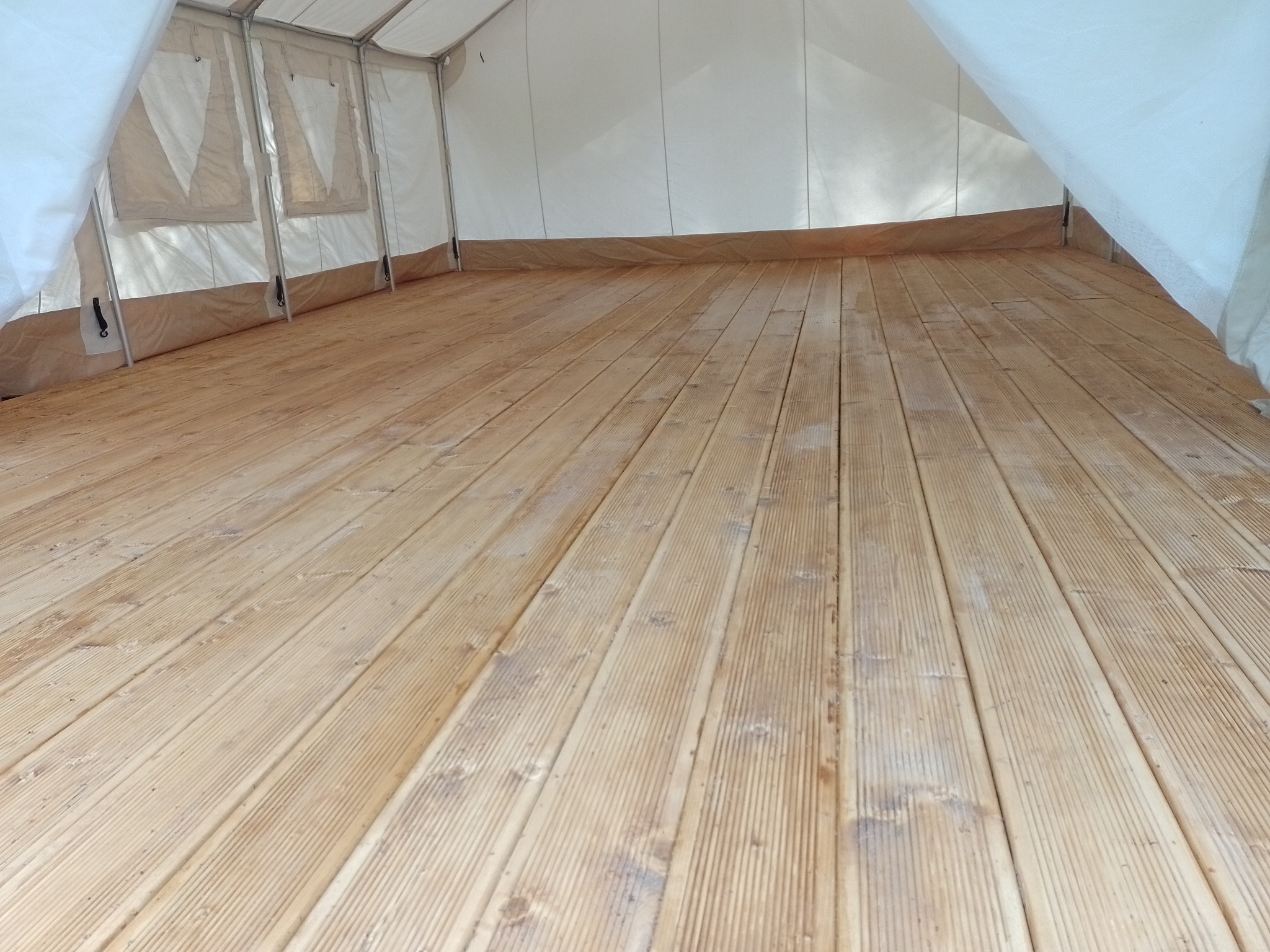 Wnętrze namiotu z drewnianą podłogą z desek o jasnym odcieniu, widoczne ślady użytkowania i naturalne sęki w drewnie, ściany namiotu wykonane z białego, półprzezroczystego materiału.