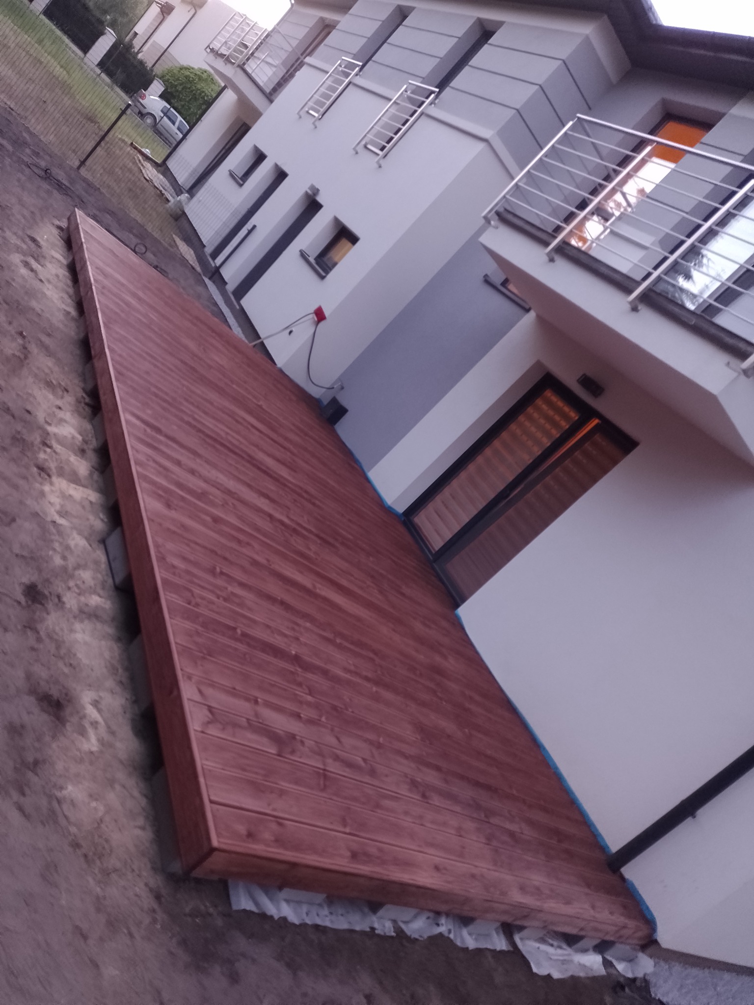 Nowo wybudowany taras z desek w odcieniach czerwieni przylegający do nowoczesnego domu z balkonem z metalową balustradą, widoczne przygotowane podłoże.