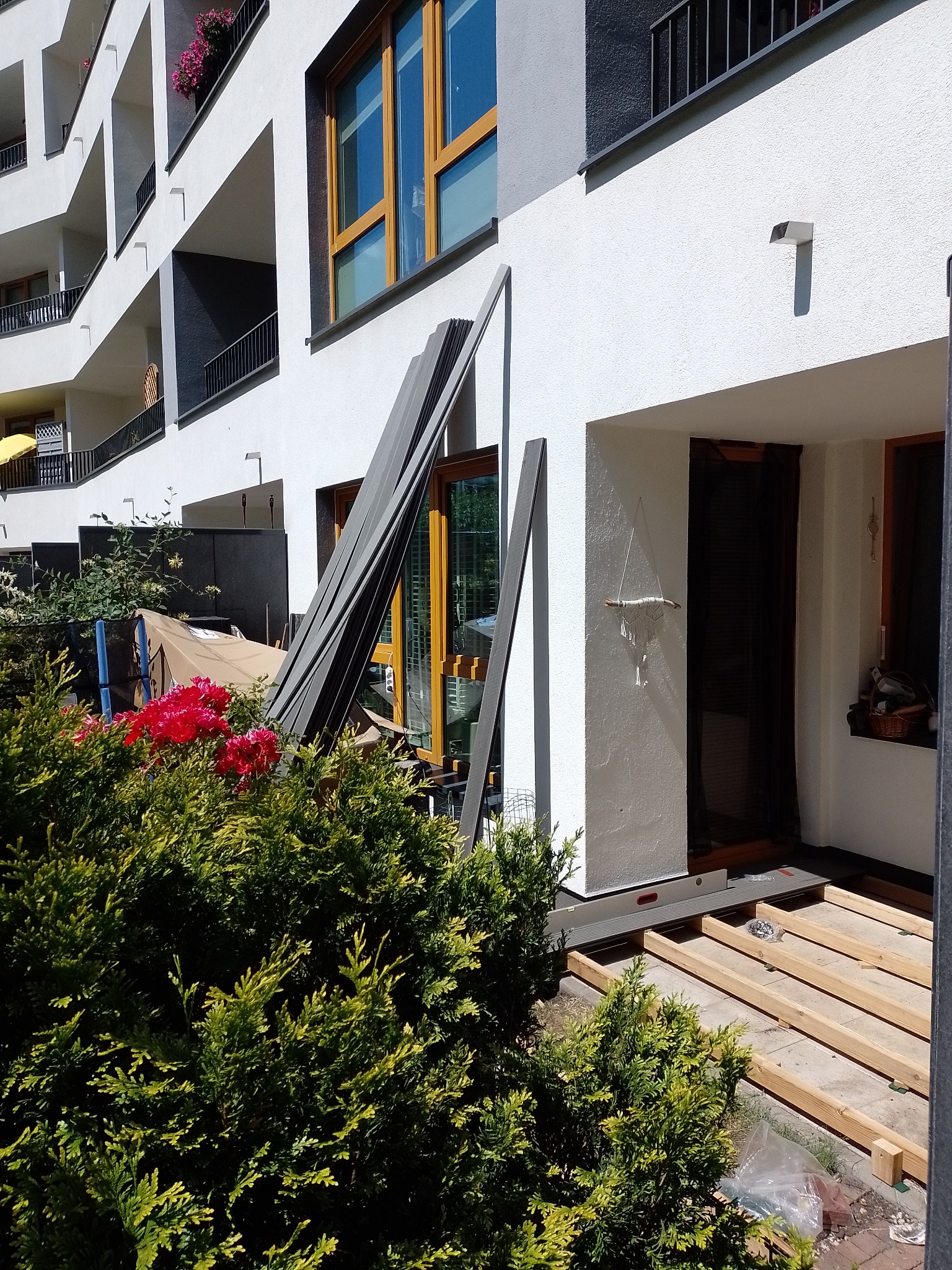 W budowie taras przy nowoczesnym apartamentowcu: drewniane legary ułożone na betonowej powierzchni, obok leżą panele kompozytowe, poziomica i worek ze śrubami.