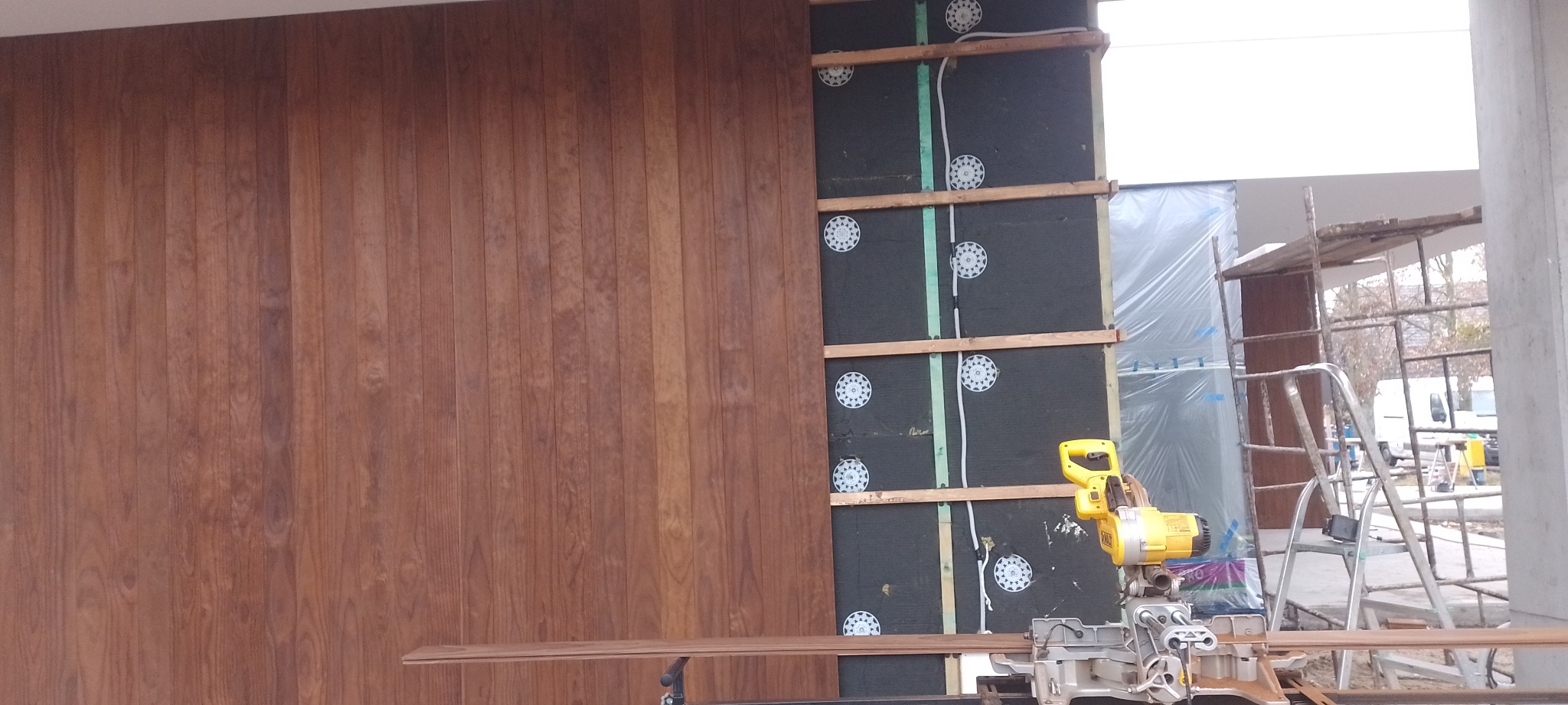 Montaż drewnianej okładziny elewacyjnej: widoczna część już zamontowana, izolacja termiczna ściany, poziomica laserowa, piła ukośnica i rusztowanie w tle.