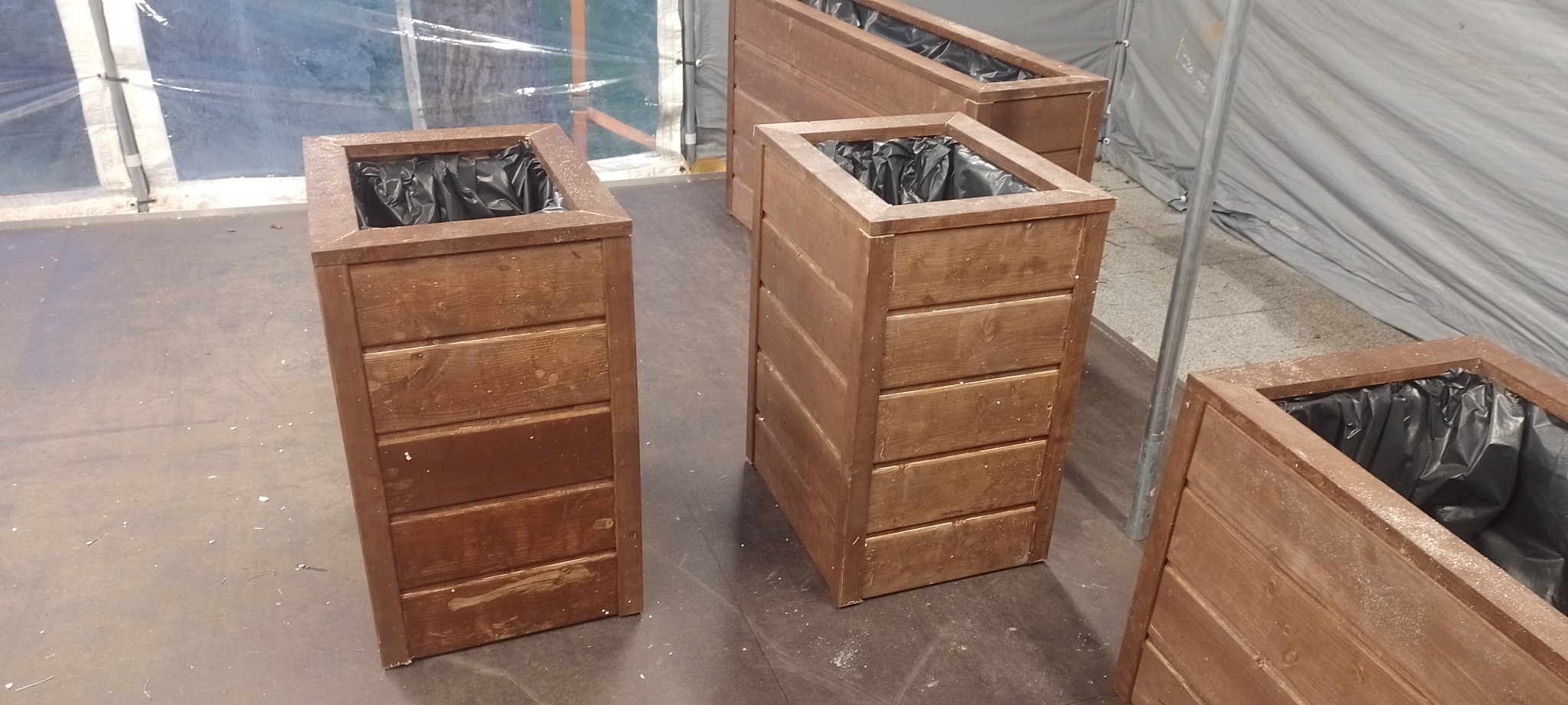 Drewniane skrzynie na rośliny z czarnym wkładem foliowym, ustawione na szarym podłożu wewnątrz namiotu.