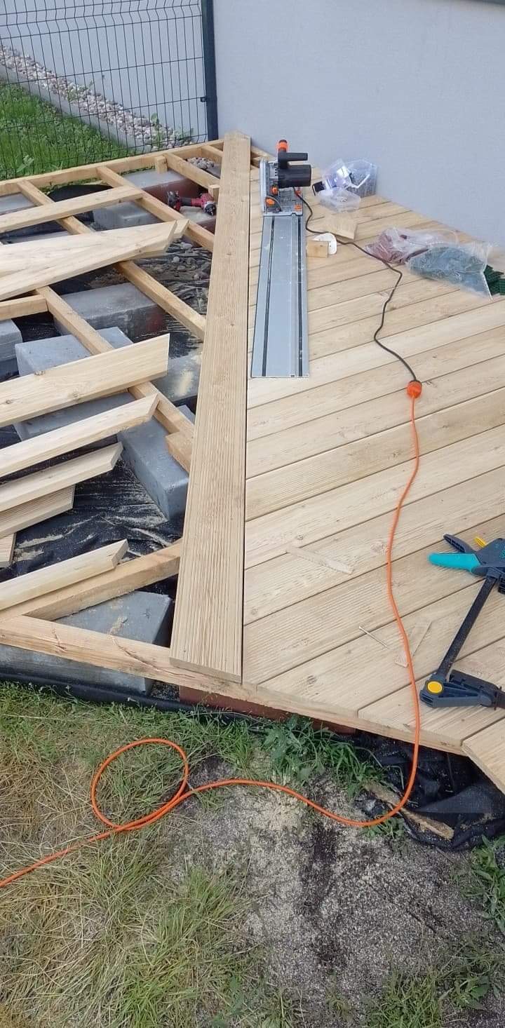 Montaż drewnianego tarasu: fragment ułożonych desek, widoczna konstrukcja nośna z bloczków betonowych, narzędzia pracy - piła tarczowa z szyną prowadzącą, zacisk stolarski i przewód zasilający.