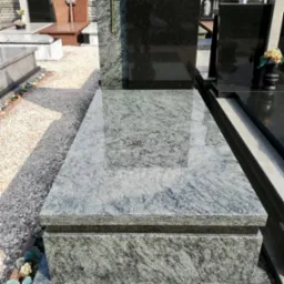 Granitowy pomnik z krzyżem w złotym kolorze, otoczony żwirowym podłożem na cmentarzu, widoczne inne nagrobki w tle.