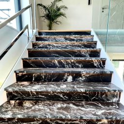 Abikam Halina Wilczyńska - Eleganckie schody wewnętrzne z czarnego kamienia z białymi żyłami, podświetlone od spodu, z metalową balustradą i rośliną w tle.