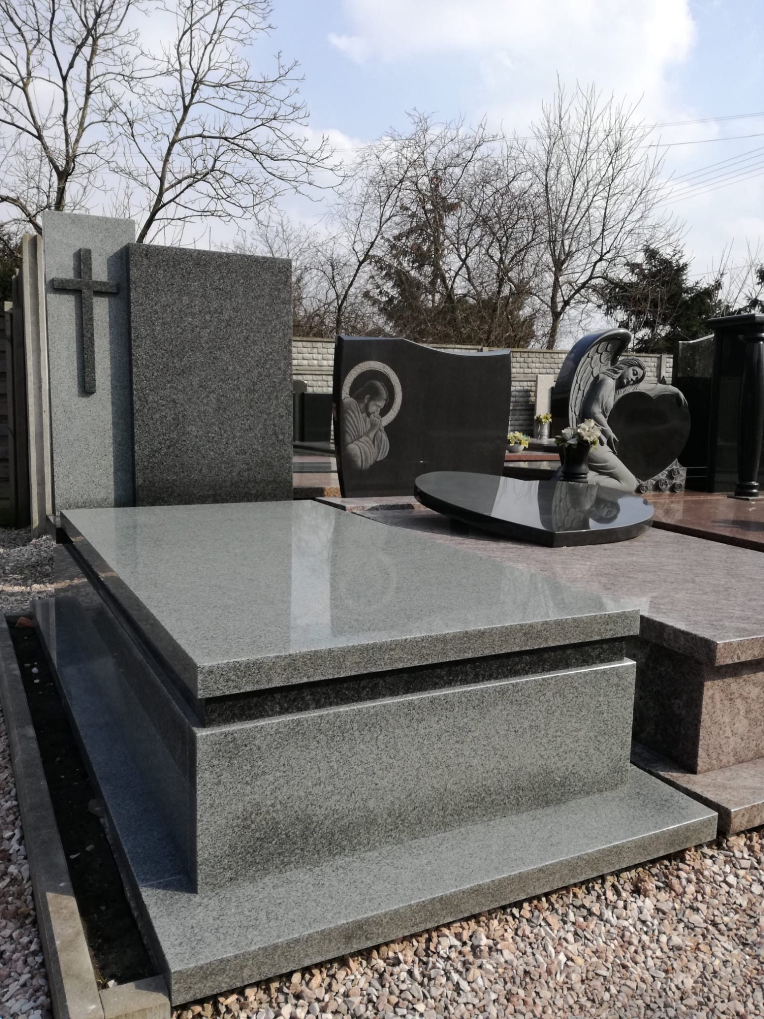 Jasnoszary, granitowy pomnik nagrobny z krzyżem, widoczny na tle innych nagrobków i drzew bez liści.