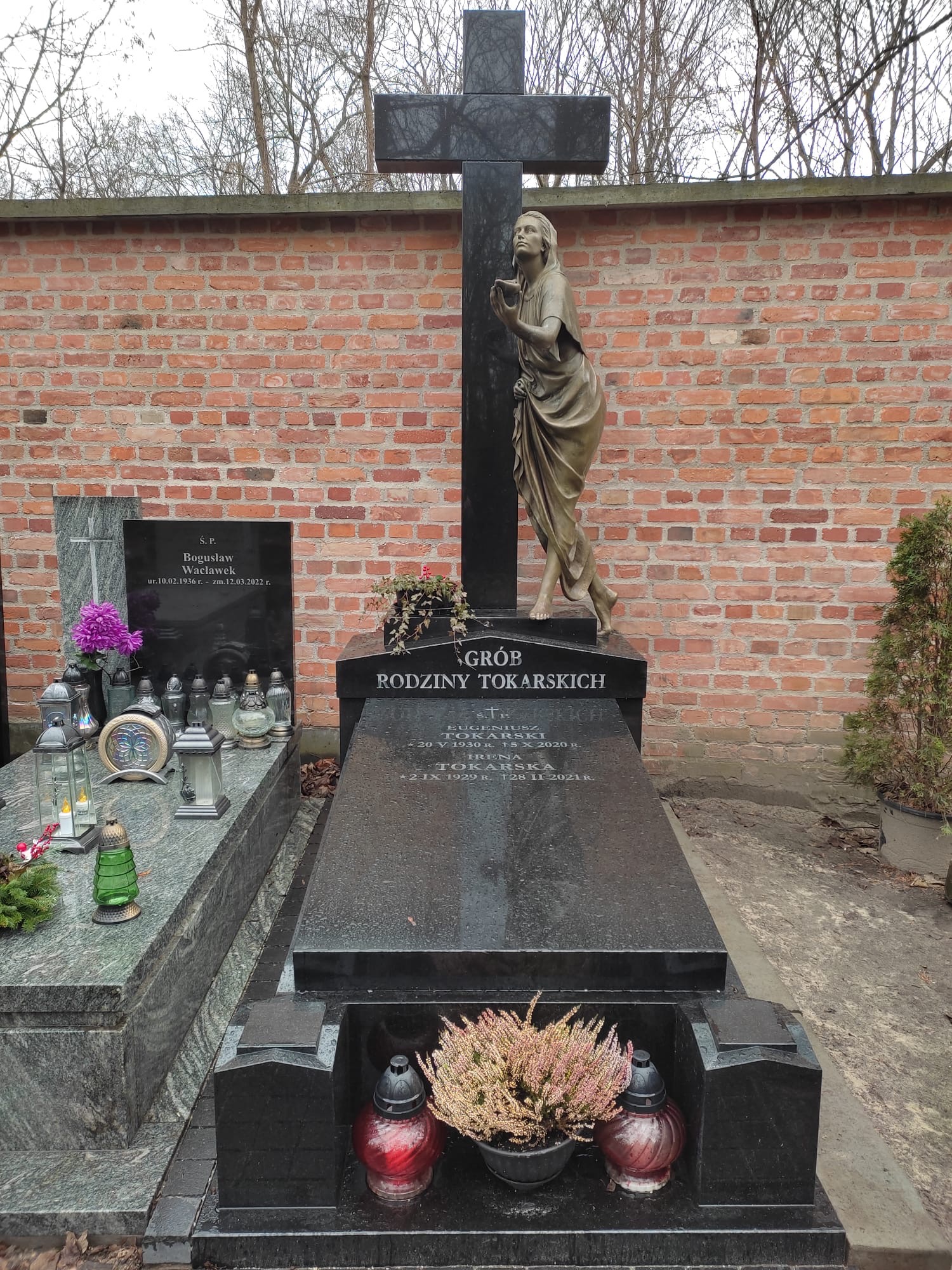 Czarny, granitowy nagrobek rodzinny z rzeźbą kobiety pod krzyżem, na cmentarzu, na tle ceglanego muru i drzew bez liści.