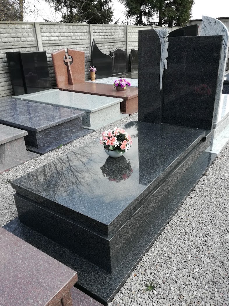 Ekspozycja różnych wzorów pomników z ciemnego granitu i jasnego kamienia w Kątach, z dekoracyjnymi kwiatami w wazonach, na żwirowym podłożu i tle betonowego ogrodzenia.