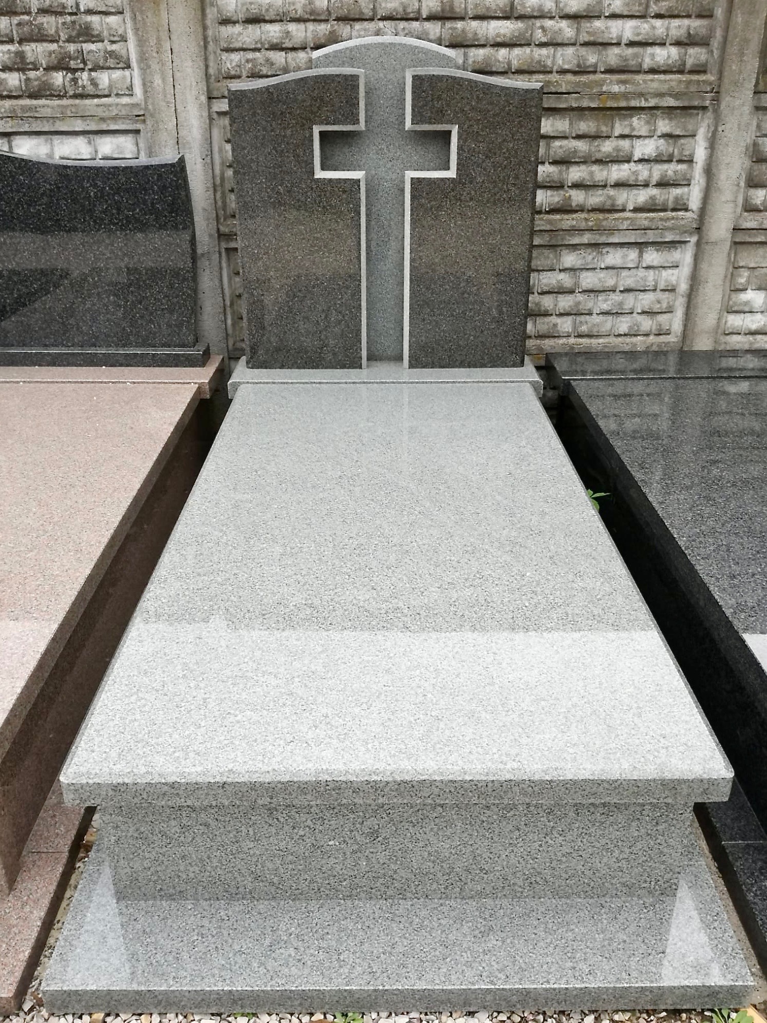 Jasnoszary, gotowy nagrobek z krzyżem na tle betonowego muru, widoczne inne nagrobki obok.