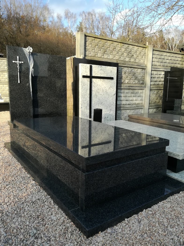 Czarny, polerowany pomnik z krzyżem i draperią, odbijający w swojej powierzchni otoczenie cmentarza, na tle betonowego muru i drzew bez liści.