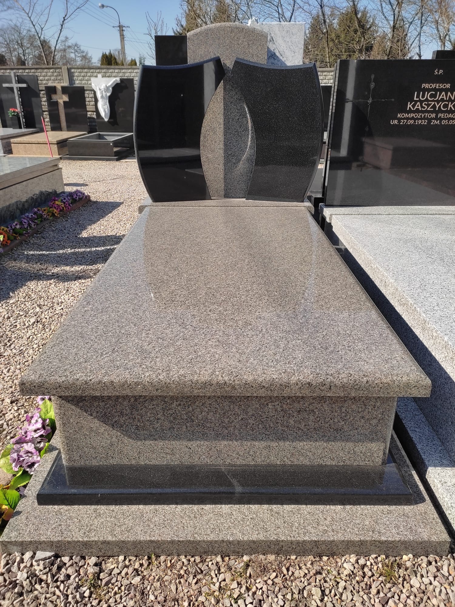 Granitowy pomnik z czarnymi tablicami na cmentarzu, widoczne kwiaty i żwir.