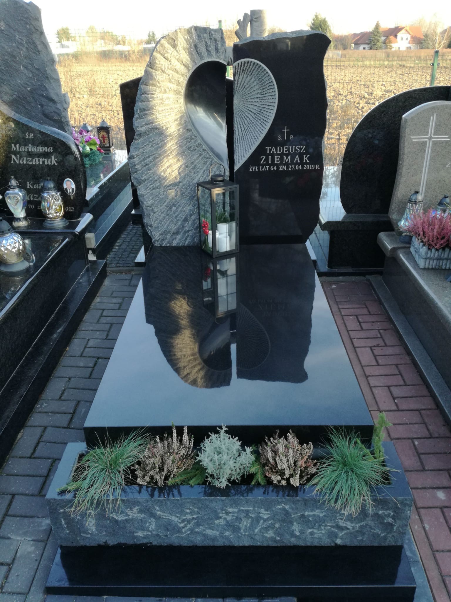 Czarny, polerowany pomnik z jasnoszarym, rzeźbionym elementem w kształcie połowy serca, obok lampion ze świecami, na tle innych nagrobków i krajobrazu.