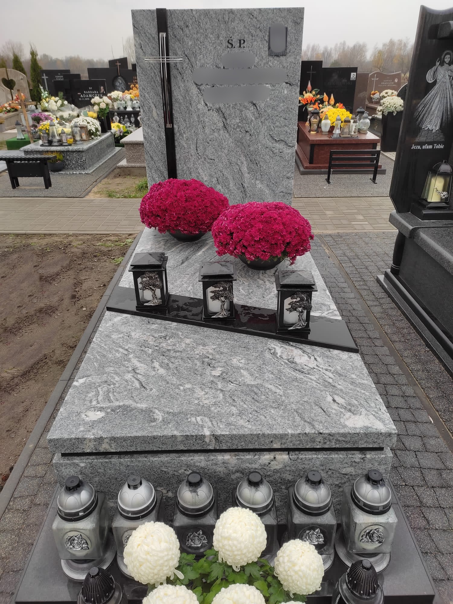 Jasnoszary granitowy pomnik z ciemnoszarym akcentem, ozdobiony czerwonymi i białymi kwiatami oraz lampionami ze wzorem drzewa, na tle innych nagrobków i szarej kostki brukowej.