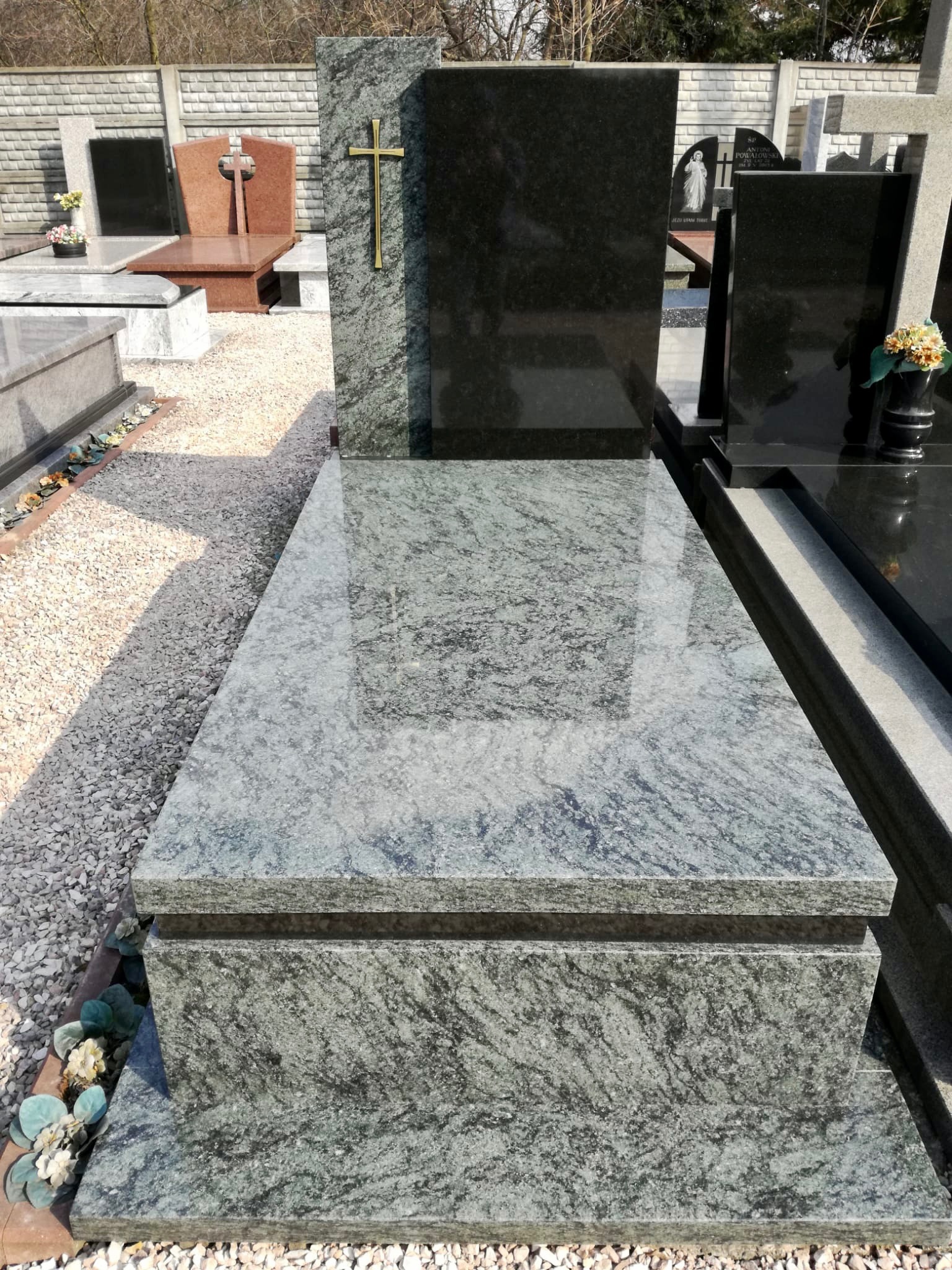 Granitowy pomnik z krzyżem w złotym kolorze, otoczony żwirowym podłożem na cmentarzu, widoczne inne nagrobki w tle.