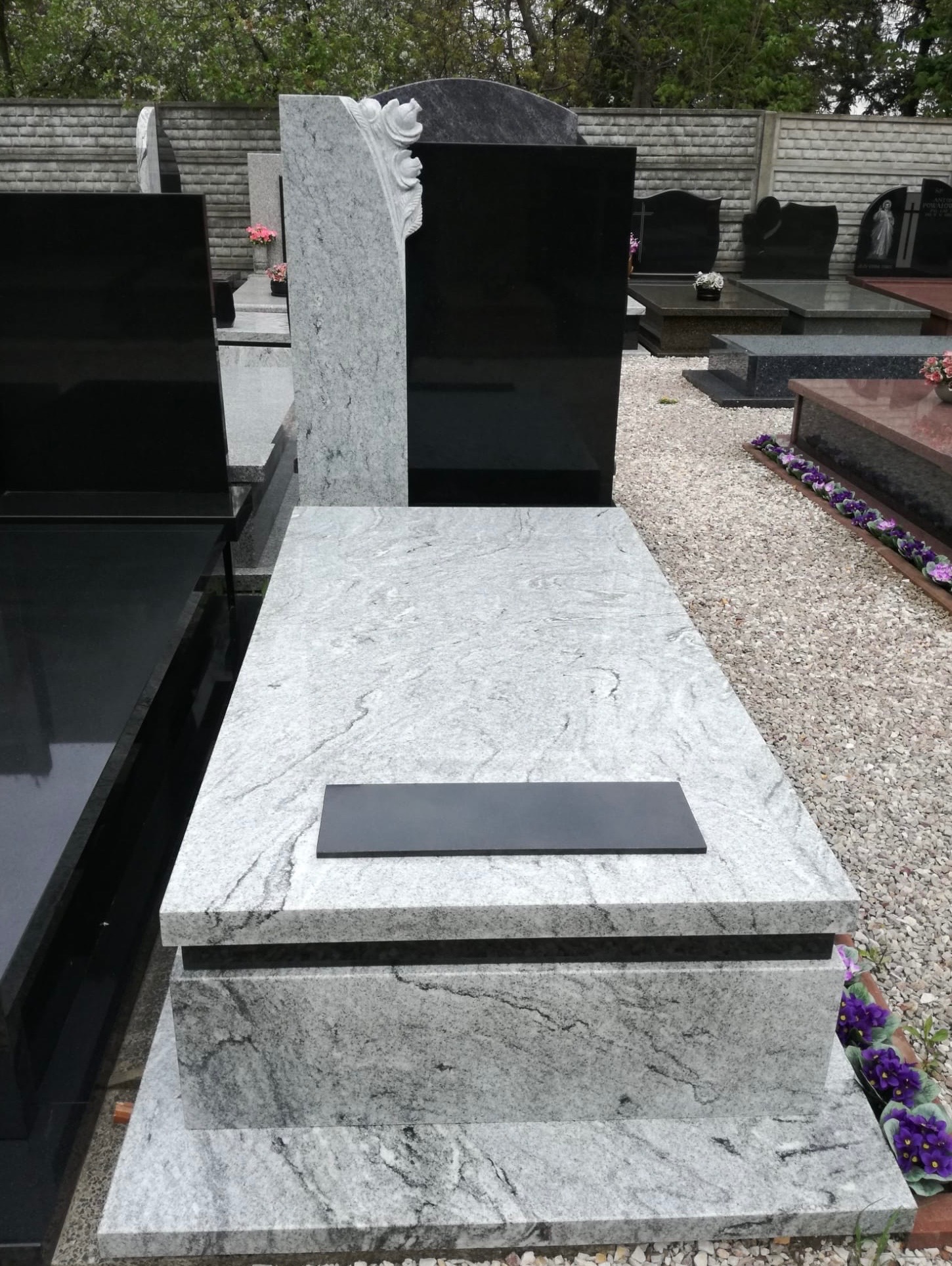 Jasnoszary, granitowy pomnik z czarną tabliczką na cmentarzu, widoczne inne nagrobki w tle.