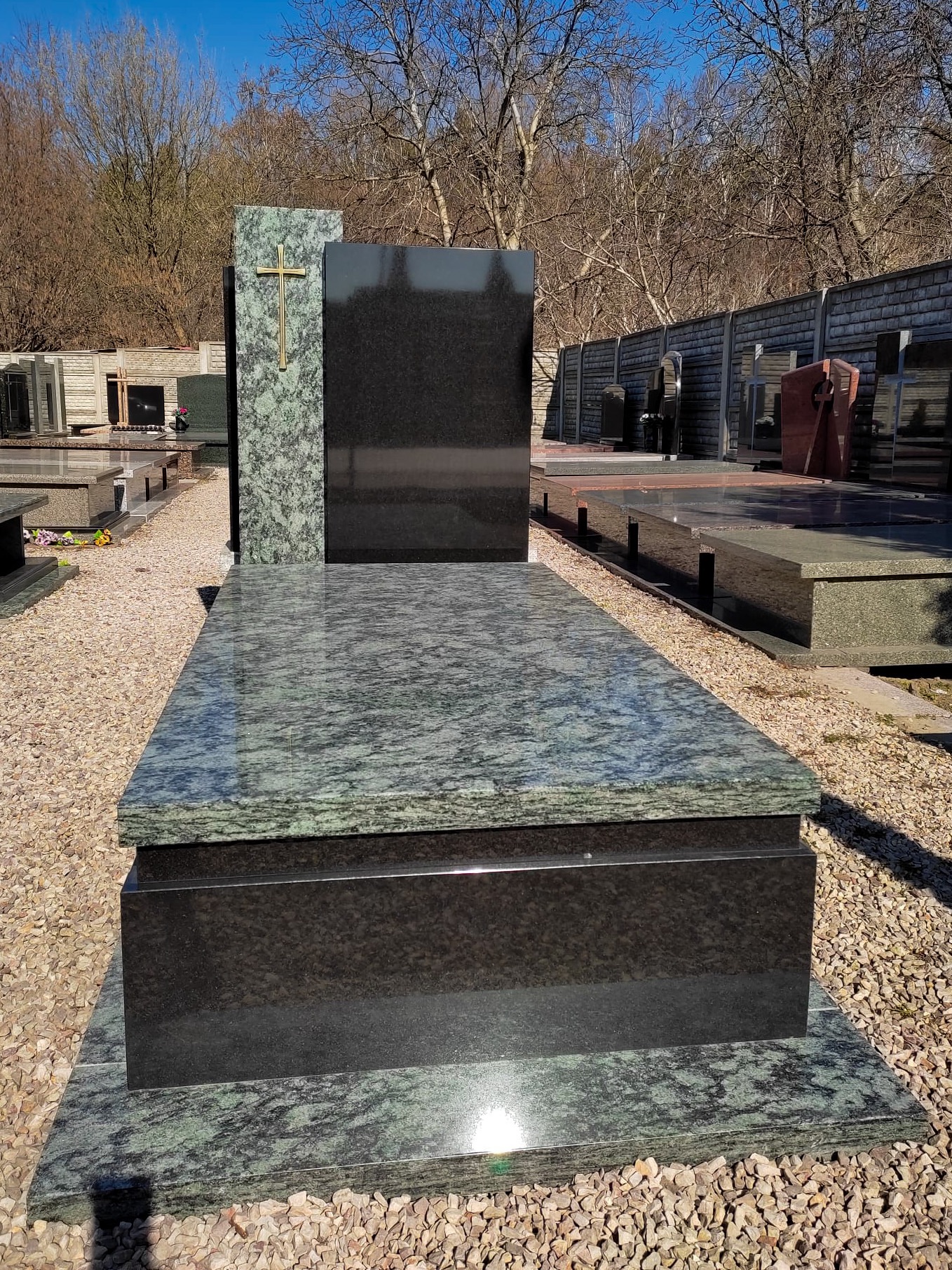 Nowoczesny, dwukolorowy pomnik z granitu z krzyżem na tle cmentarza z nagrobkami i drzewami bez liści.