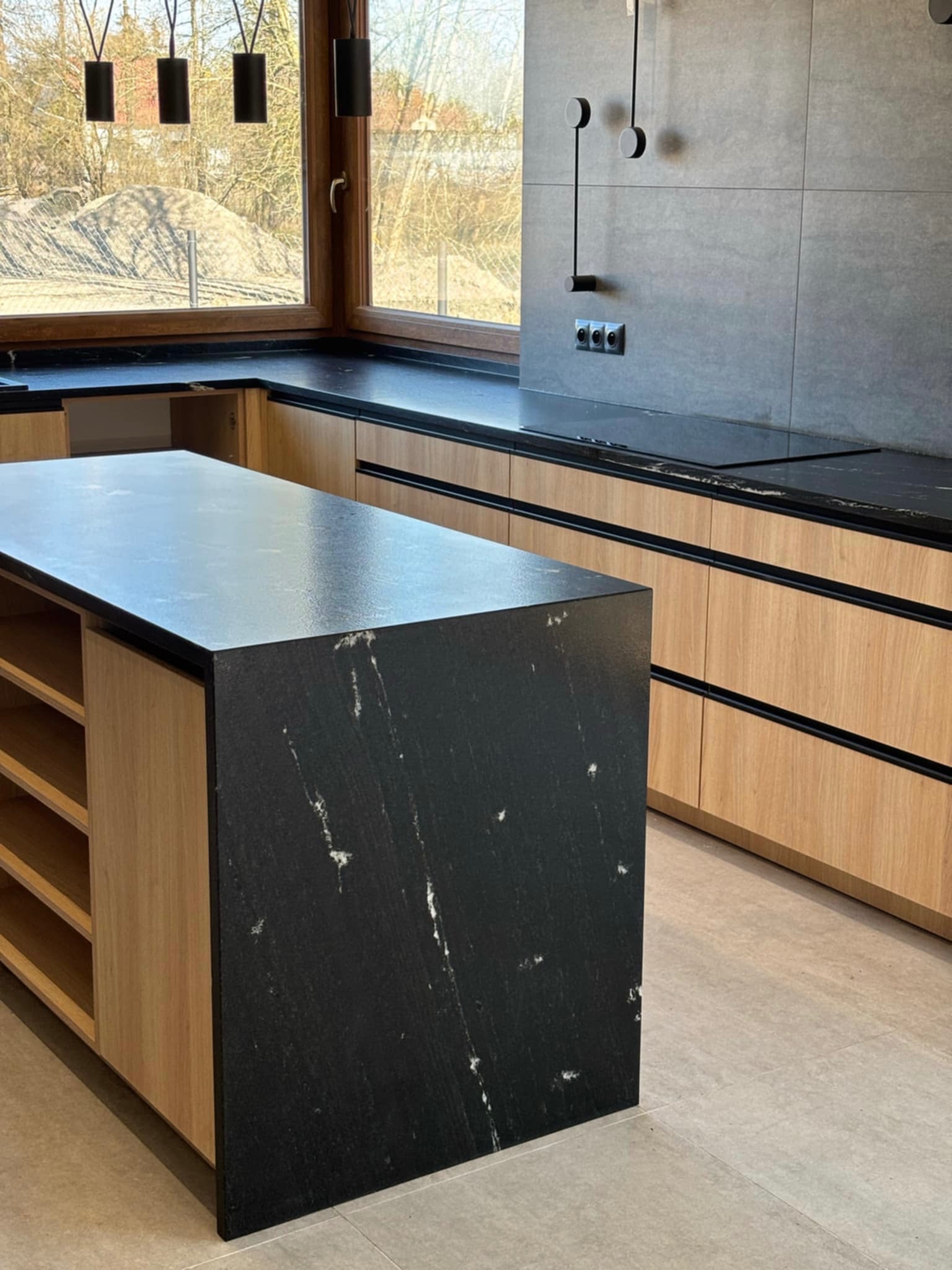 Nowoczesna kuchnia z wyspą i blatem z czarnego kamienia z białymi żyłkami, drewniane szafki, szare płytki na ścianie i minimalistyczne oświetlenie.