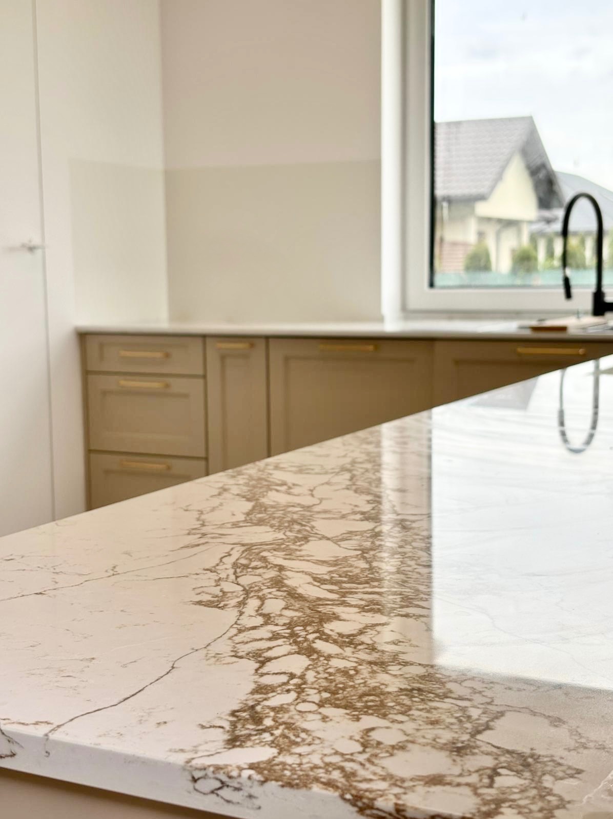 Błyszczący blat kamienny z unikalnym wzorem żyłkowania w odcieniach brązu, na tle beżowych szafek kuchennych i okna z widokiem na dom.