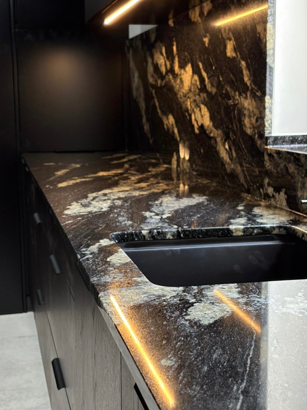 Czarny, polerowany blat kamienny z unikalnym wzorem, zintegrowany z czarnym zlewozmywakiem i oświetleniem LED, tworzący elegancką przestrzeń kuchenną.