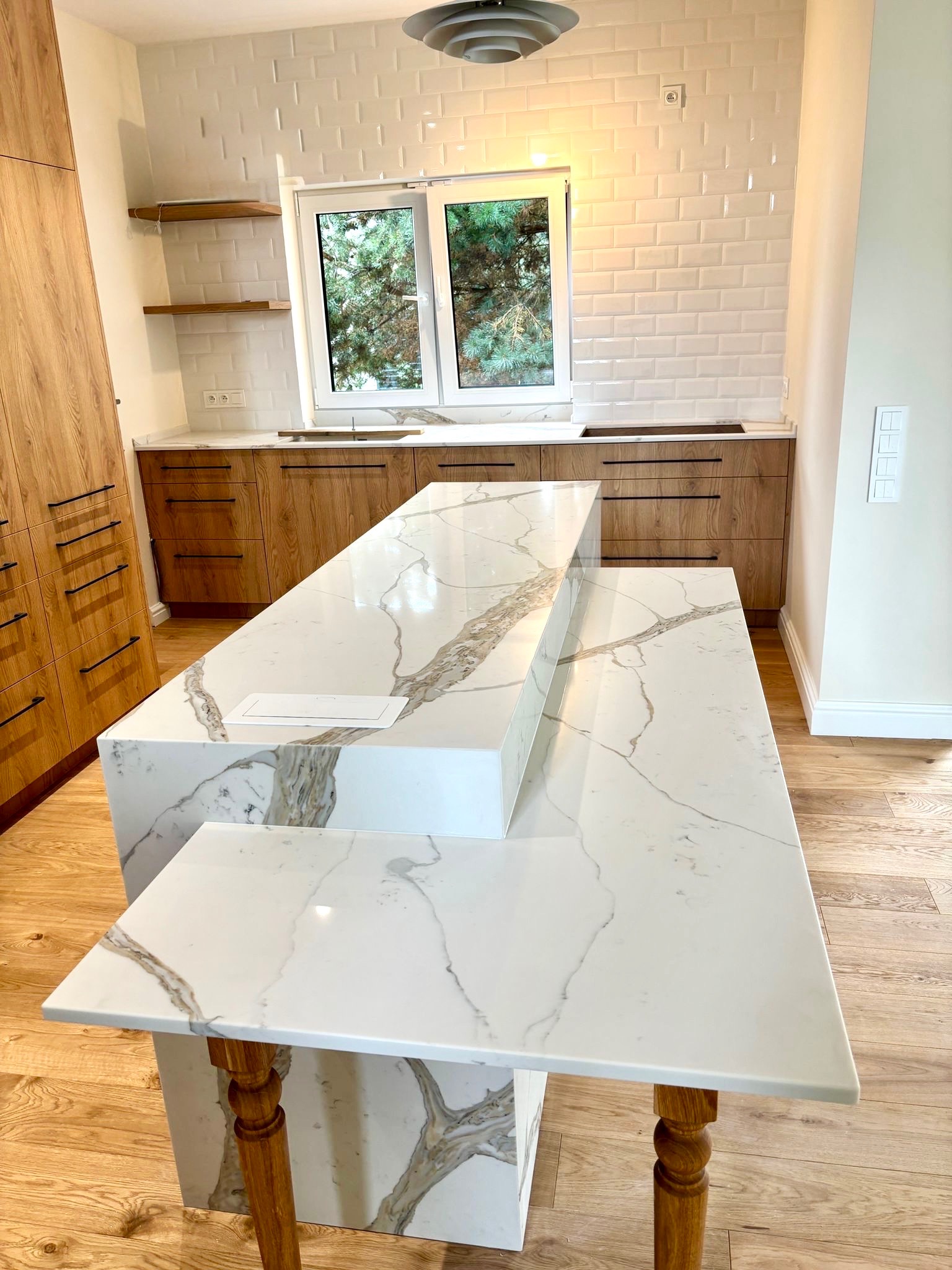 Dwupoziomowy blat kamienny w kuchni z drewnianymi szafkami i białymi płytkami. Blat z białego kamienia z szarymi żyłami, wsparty na drewnianych nogach.