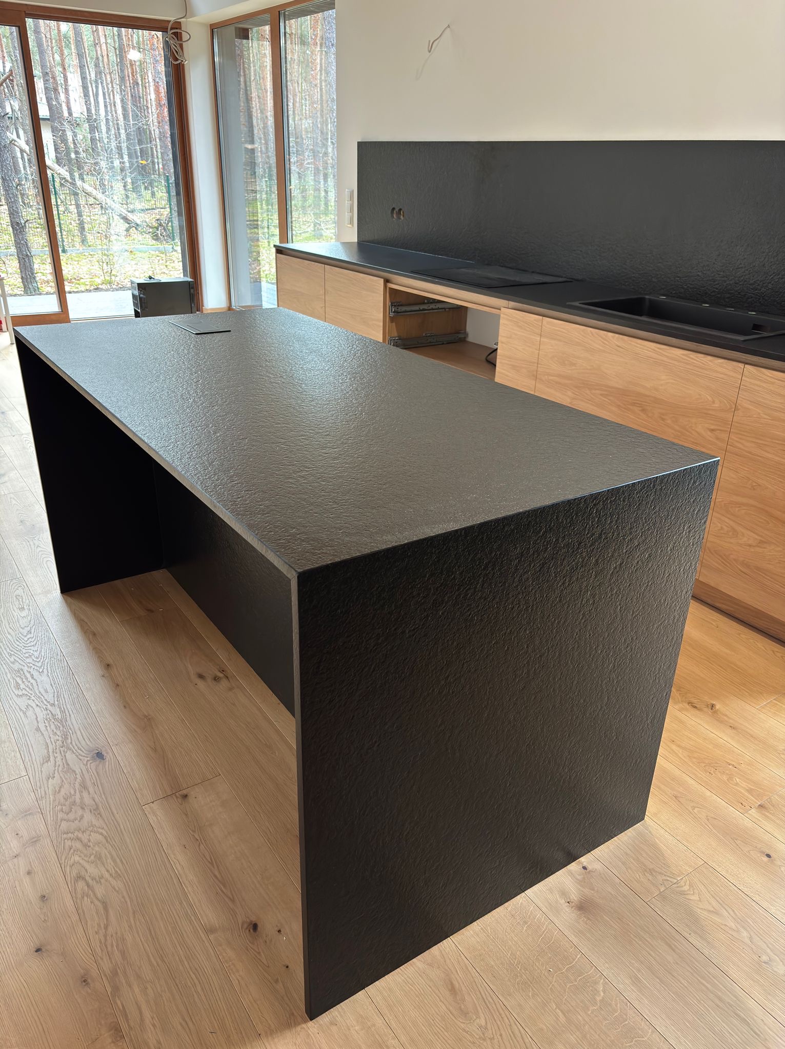 Czarny, teksturowany blat wyspowy z kamienia w nowoczesnej kuchni z drewnianymi szafkami i dużym oknem z widokiem na las. Minimalistyczny design, naturalne światło.