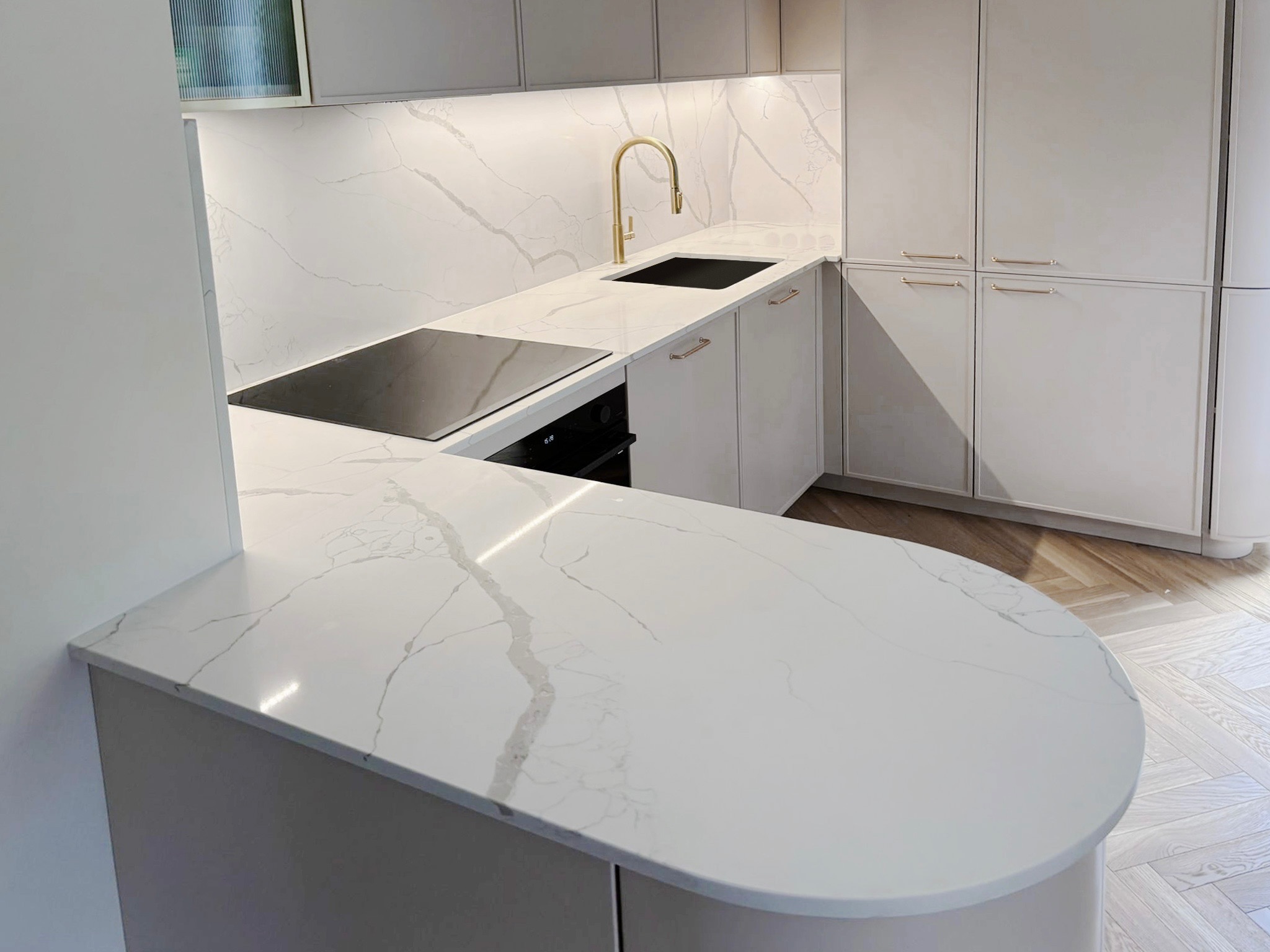 Jasna kuchnia z blatem z konglomeratu w odcieniach bieli i szarości, złota bateria, płyta indukcyjna. Uwagę przykuwa zaokrąglony kształt blatu.