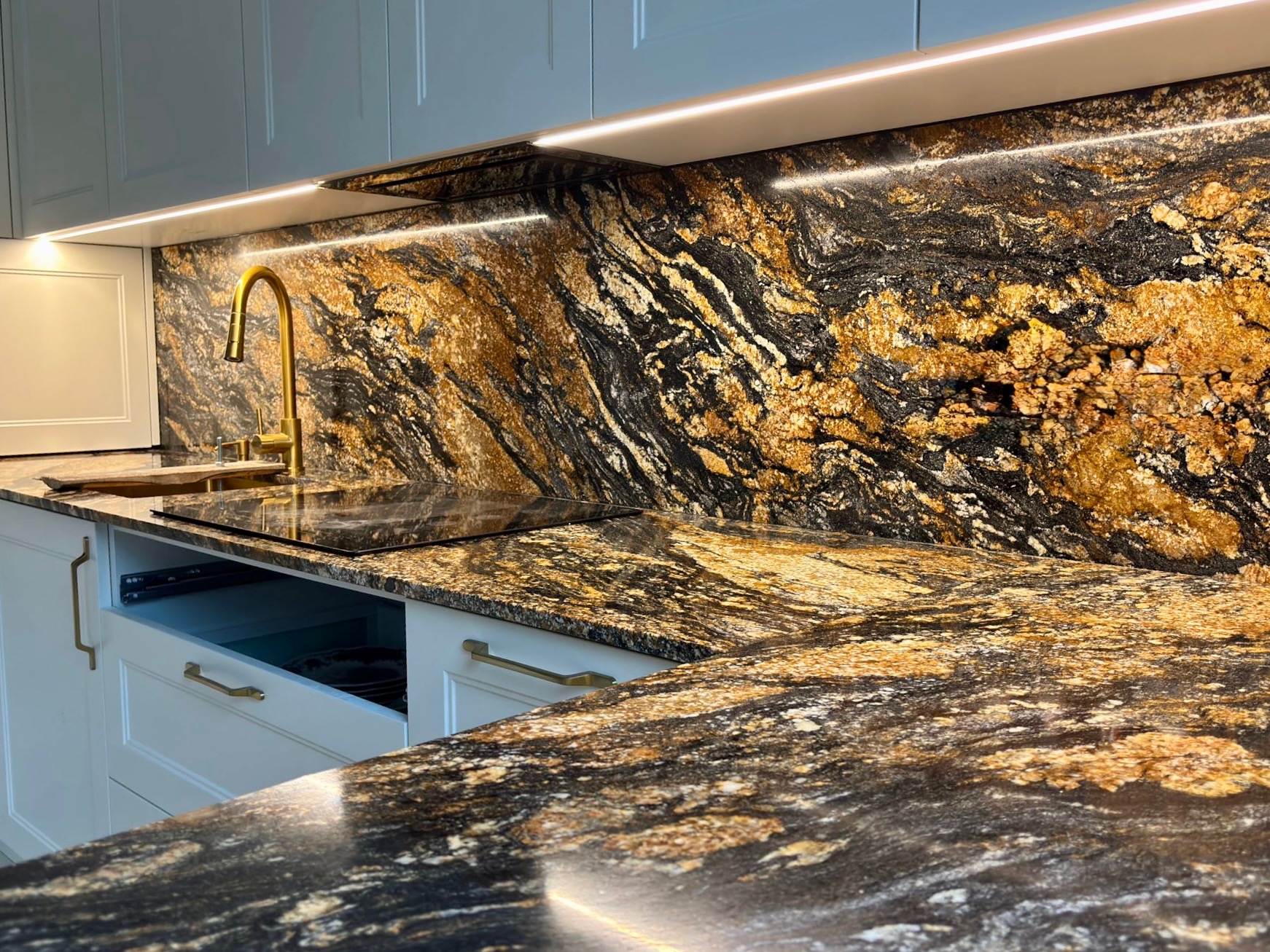 Czarno-złoty blat kamienny z połyskiem, dopasowany do ściany nad blatem, złota bateria, białe szafki z otwartą szufladą, oświetlenie LED pod szafkami.