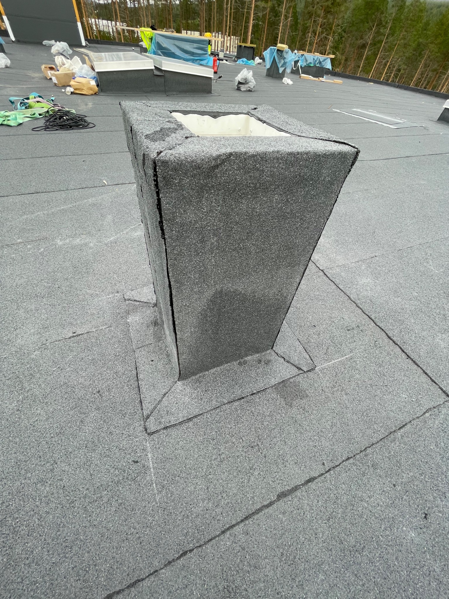 Szary, wykończony komin wentylacyjny na płaskim dachu pokrytym papą, widok z góry, widoczne obróbki blacharskie i fragmenty okien dachowych w tle.