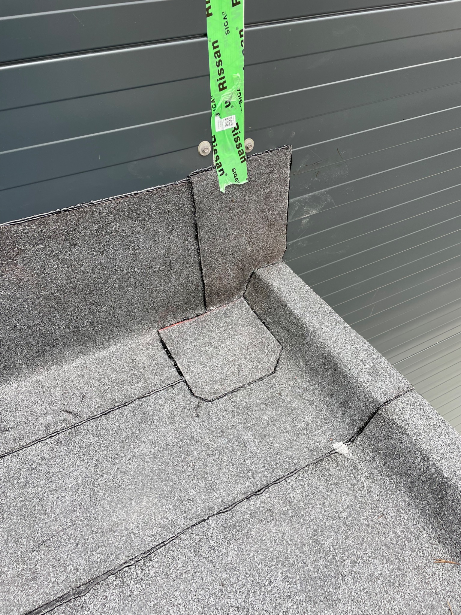 Szczegółowe ujęcie płaskiego dachu pokrytego papą, z widocznymi łączeniami i detalami wykończenia przy ścianie z blachy trapezowej, zabezpieczonej taśmą montażową.