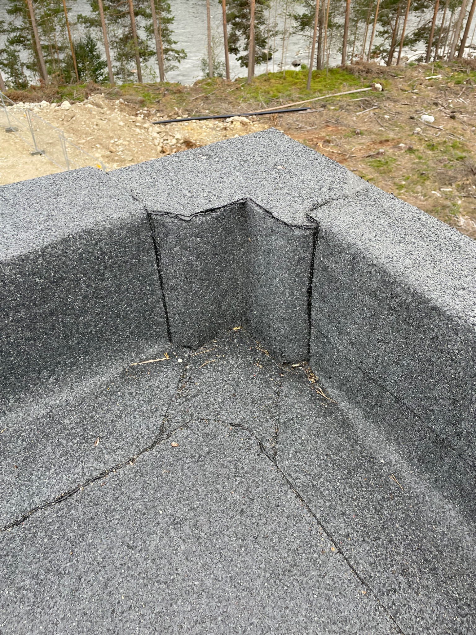 Fragment pokrycia papą termozgrzewalną na płaskim dachu z widocznymi spoinami i pęknięciem, w tle drzewa i brzeg jeziora.