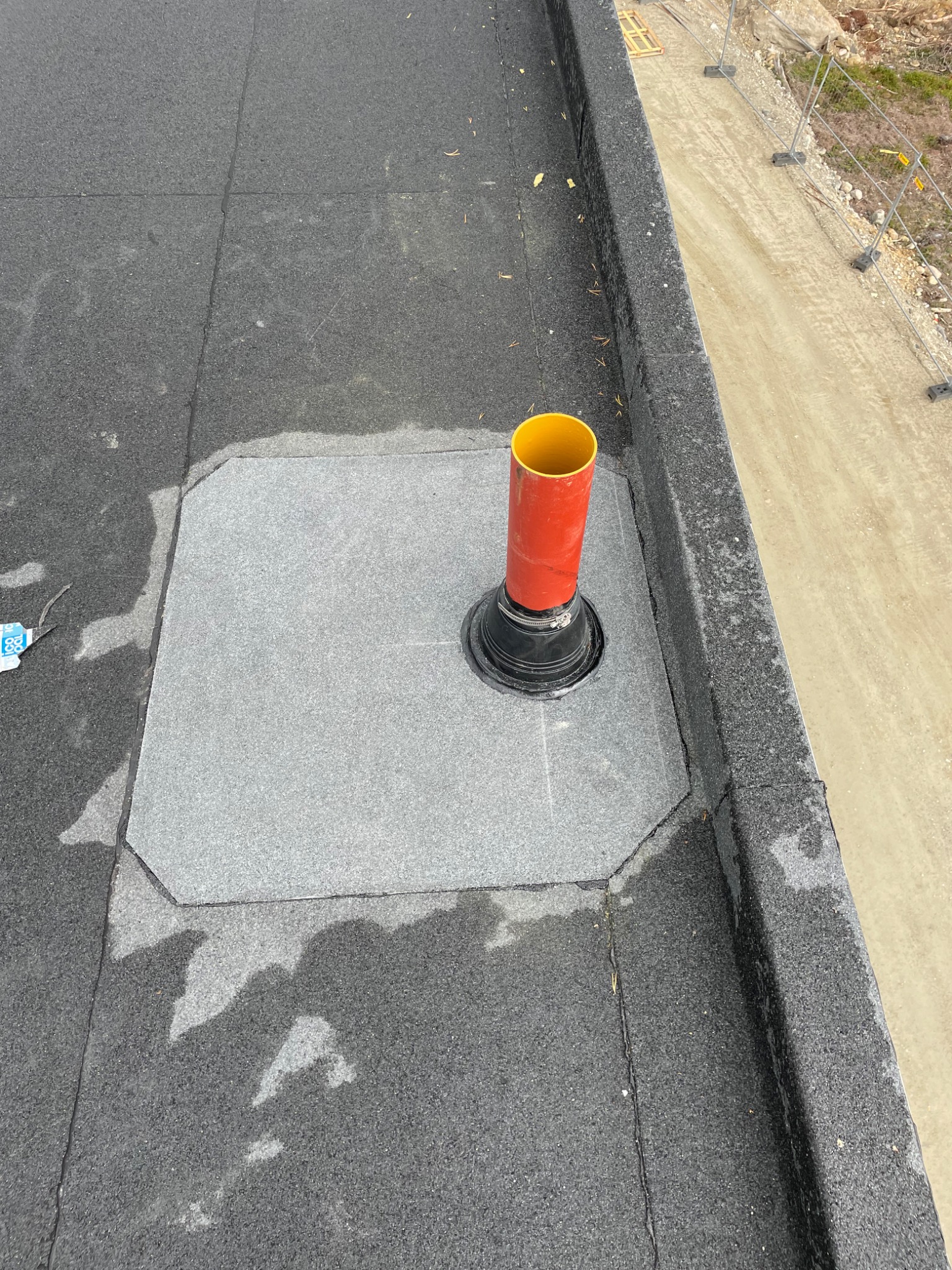 Wykończony fragment płaskiego dachu pokrytego papą z zamontowanym pionowym kominkiem wentylacyjnym w kolorze czerwono-żółtym, na tle piaszczystej drogi i barierek budowlanych.