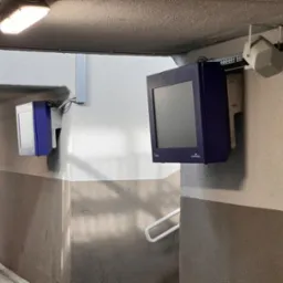 Dwa ekrany informacyjne w fioletowej obudowie zamontowane na ścianie korytarza, obok głośnik na szynie, widoczny numer platformy '2'.