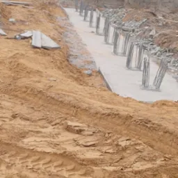 Wykop pod fundament z wylanym betonem i wystającymi prętami zbrojeniowymi, widoczne ślady opon ciężkiego sprzętu w piasku.