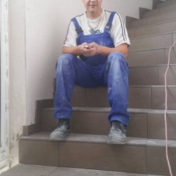 Kroman - Pracownik w niebieskim kombinezonie siedzi na schodach wyłożonych brązowymi płytkami, trzymając coś w dłoniach. Ściana za nim jest surowa, a obok widoczna rama okienna.