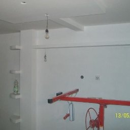 Kroman - Wnętrze po świeżym malowaniu ścian i sufitu z widocznym podnośnikiem do płyt gipsowo-kartonowych, wiszącą żarówką bez klosza i półkami w ścianie, z butelką wody na jednej z nich.