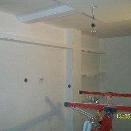 Kroman - Wnętrze w trakcie malowania na biało, widoczne niedokończone sufity podwieszane, gołe żarówki zwisające z sufitu i czerwony stojak malarski.