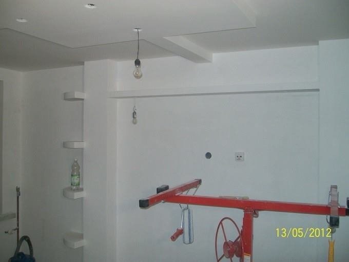 Wnętrze po świeżym malowaniu ścian i sufitu z widocznym podnośnikiem do płyt gipsowo-kartonowych, wiszącą żarówką bez klosza i półkami w ścianie, z butelką wody na jednej z nich.