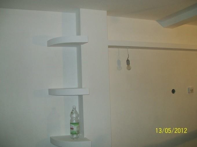 Białe, półokrągłe półki narożne na białej ścianie, z butelką wody na najniższej półce. Widoczne niedokończone elementy instalacji elektrycznej.