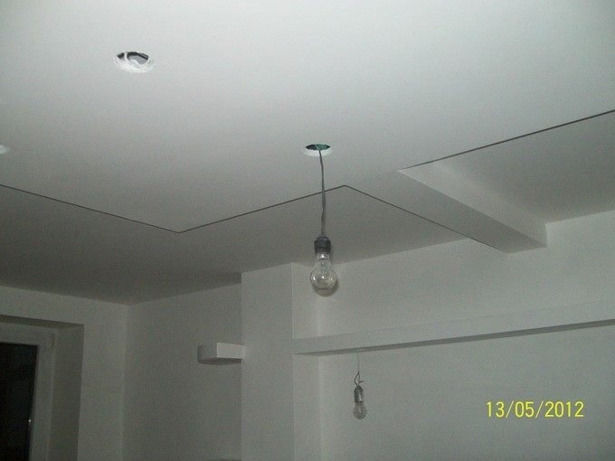 Biały sufit z geometrycznymi wzorami i wystającymi elementami, z widocznymi kablami i żarówkami wiszącymi z otworów, stanowiący etap prac wykończeniowych.