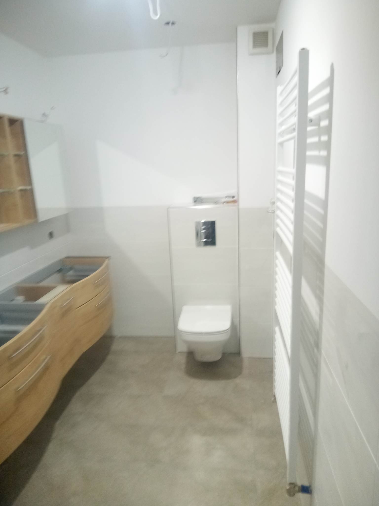 Wnętrze łazienki w trakcie prac wykończeniowych: zamontowana toaleta podtynkowa, grzejnik drabinkowy, częściowo zamontowana szafka pod umywalkę z drewnianym frontem, płytki na ścianach do połowy...