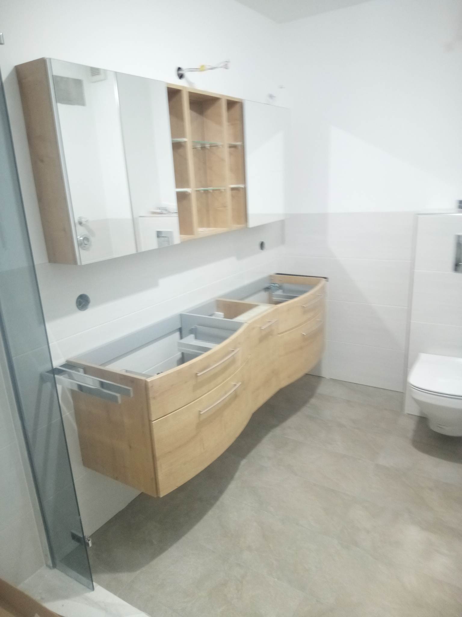 Niedokończona, podwieszana szafka łazienkowa z drewnianymi frontami i szufladami, zamontowana pod lustrem z szafką wiszącą z półkami, widoczna toaleta i fragment szklanej kabiny prysznicowej.